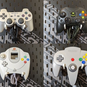 May include: Four different video game controllers mounted on a pegboard wall. The controllers are from the Playstation, Xbox, Nintendo 64, and Sega Dreamcast consoles. The controllers are all gray and white except for the Xbox controller, which is black and green.