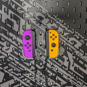 May include: Two Nintendo Switch Joy-Con controllers, one purple and one yellow, are hanging on a black pegboard wall.