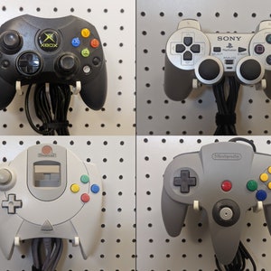 May include: Four video game controllers: an Xbox controller, a PlayStation controller, a Dreamcast controller, and a Nintendo 64 controller. Each controller is mounted on a white pegboard. The Xbox controller is black, the PlayStation controller is gray, the Dreamcast controller is white, and the Nintendo 64 controller is gray.