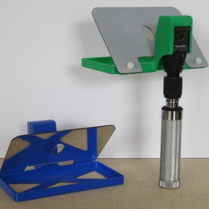 Op de afbeelding: Twee ophthalmoscoop houders, een groene en een blauwe, met een spiegel en een gat voor de ophthalmoscoop. De groene houder is aan de muur bevestigd en de blauwe houder staat op een tafel.