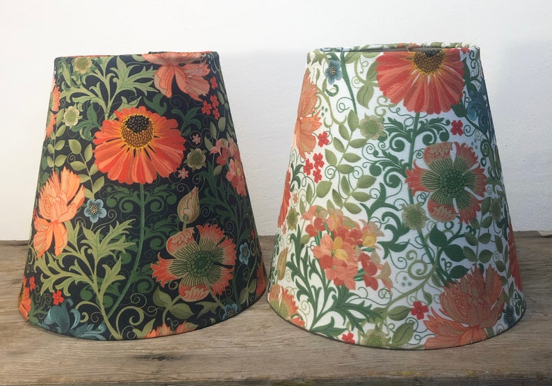 May include: Two lampshades with floral patterns. One shade has a dark background with orange and green flowers. The other shade has a white background with orange and green flowers. Both shades have a conical shape.