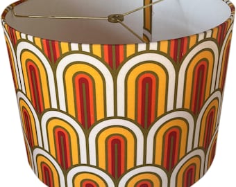 May include: A cylindrical lampshade with a retro design. The shade features a repeating pattern of arched shapes in shades of yellow, orange, red, and white, with a gold-colored metal frame. The interior is white.