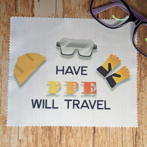 May include: A white cloth with the text "HAVE PPE WILL TRAVEL" and illustrations of safety equipment, including a hard hat, safety glasses, and gloves. Black and purple eyeglasses rest on the cloth.