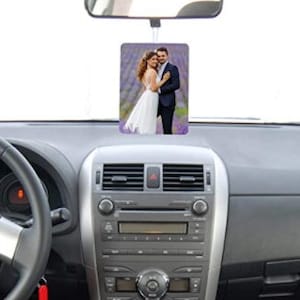 May include: Rectangular car air freshener featuring a wedding photo, suspended from a car's rearview mirror. The car's dashboard is visible, including the steering wheel and central console.