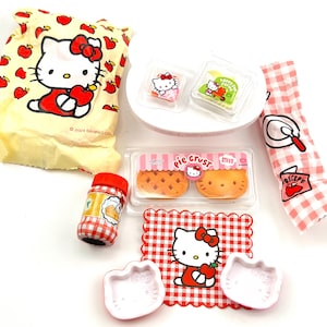 May include: A Hello Kitty themed play food set with a red and white checkered tablecloth, a bag of apples, a jar of pie filling, two pie crusts, a plate of pie filling, and two pie molds.