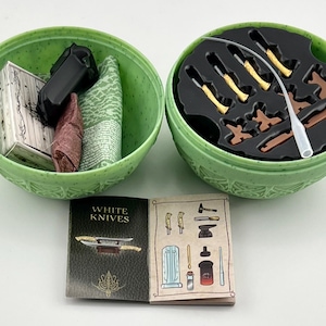 May include: Two green plastic containers with lids, one open and one closed. The open container holds a small book with the words "White Knives" on the cover, a small brown and red rolled up piece of fabric, a small black object, and a small white box. The closed container has a black interior with small brown and gold objects and a white tube.