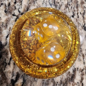May include: Two yellow dice with white dots are encased in a clear resin coaster with gold glitter. The coaster is round and has a gold rim.