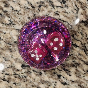 May include: Two pink dice with white dots are inside a clear, round, pink glitter-filled container.