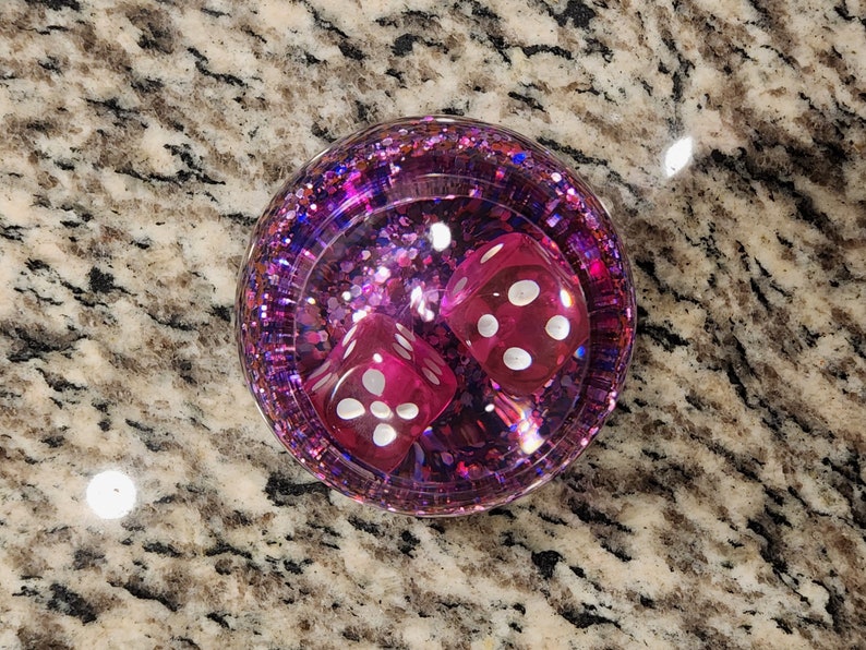 May include: Two pink dice with white dots are inside a clear, round, pink glitter-filled container.