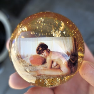 May include: A clear glass paperweight with gold flecks and a picture of a woman in a vintage style setting. The woman is wearing a white bra and is sitting on a bed with a pillow.