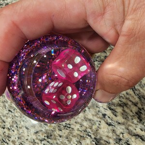 May include: A clear sphere filled with pink glitter and two pink dice with white dots. The dice are visible through the clear sphere.