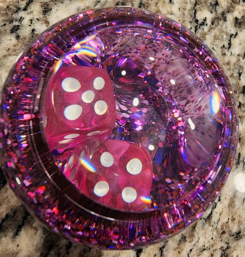 May include: Two pink dice with white dots are inside a clear, round, pink and purple glitter-filled container.