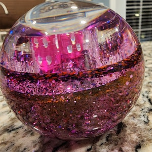 May include: A clear glass sphere filled with pink and orange glitter. The glitter is suspended in a clear liquid and moves around when the sphere is shaken.