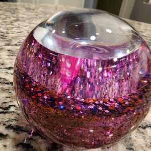 May include: A clear glass sphere filled with pink, red, and blue glitter. The glitter is suspended in a clear liquid.