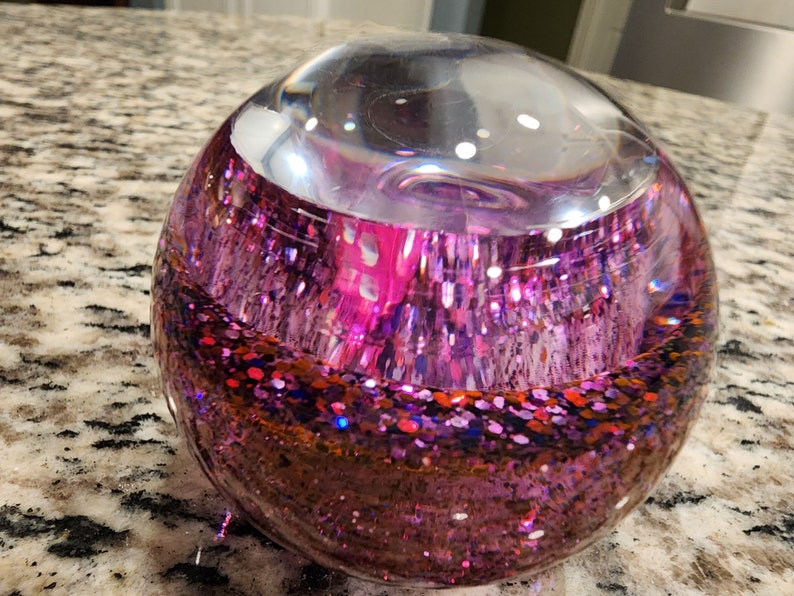 May include: A clear glass sphere filled with pink, red, and blue glitter. The glitter is suspended in a clear liquid.
