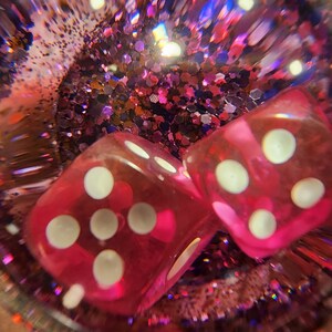 May include: Two pink dice with white dots are inside a clear sphere filled with pink and purple glitter.