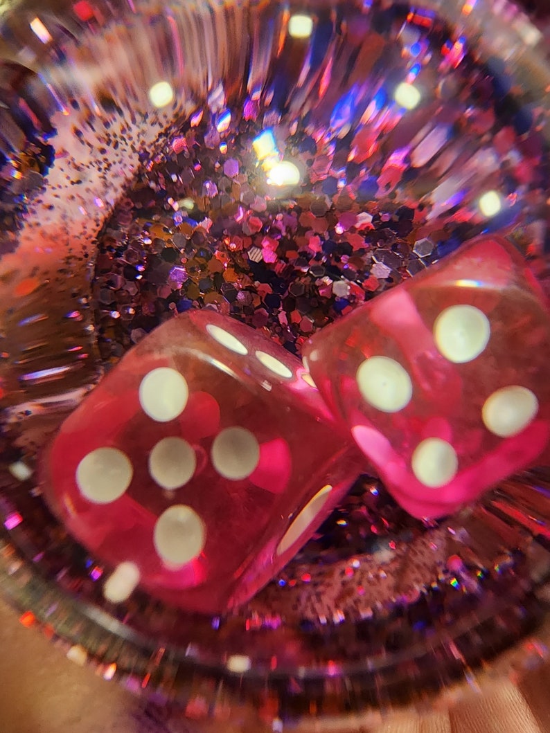 May include: Two pink dice with white dots are inside a clear sphere filled with pink and purple glitter.