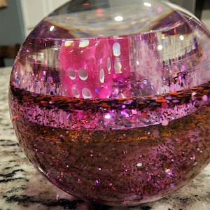 May include: A clear glass sphere filled with pink glitter and a pink die. The die is partially submerged in the glitter.