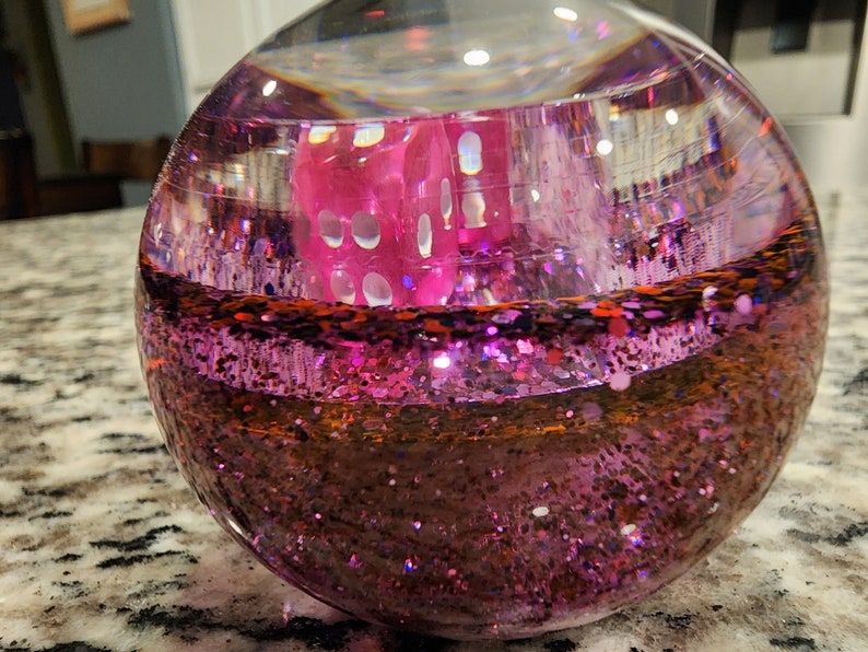 May include: A clear glass sphere filled with pink glitter and a pink die. The die is partially submerged in the glitter.