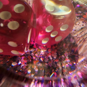 May include: Two red dice with white dots are partially submerged in a clear container filled with pink, gold, and purple glitter. The dice are slightly blurred due to the glitter.