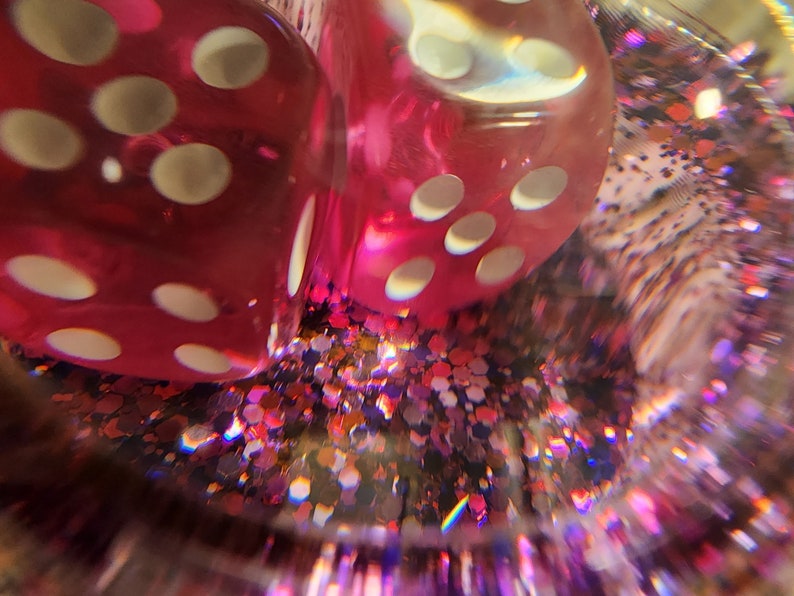 May include: Two red dice with white dots are partially submerged in a clear container filled with pink, gold, and purple glitter. The dice are slightly blurred due to the glitter.
