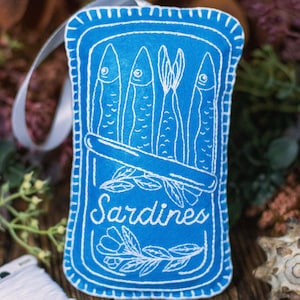 May include: A blue felt ornament shaped like a can of sardines. The can has white embroidery of sardines and the word "Sardines".