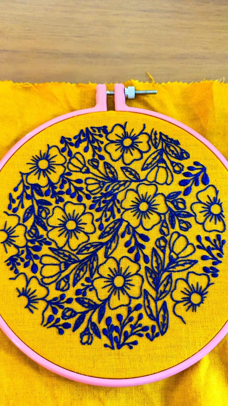 Puede incluir: Primer plano de un bastidor de bordado con un dise&ntilde;o floral. La tela amarilla est&aacute; bordada con flores y hojas azules. El bastidor es rosa y el fondo es tela amarilla.