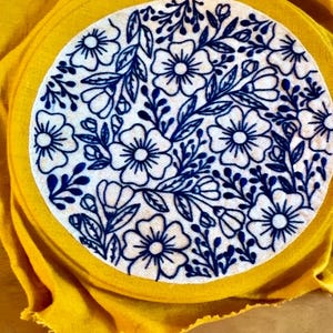 Puede incluir: Un c&iacute;rculo de tela blanca con un dise&ntilde;o de bordado floral azul, sobre un fondo de tela amarilla. El bordado presenta flores y hojas detalladas. La tela amarilla rodea el c&iacute;rculo.