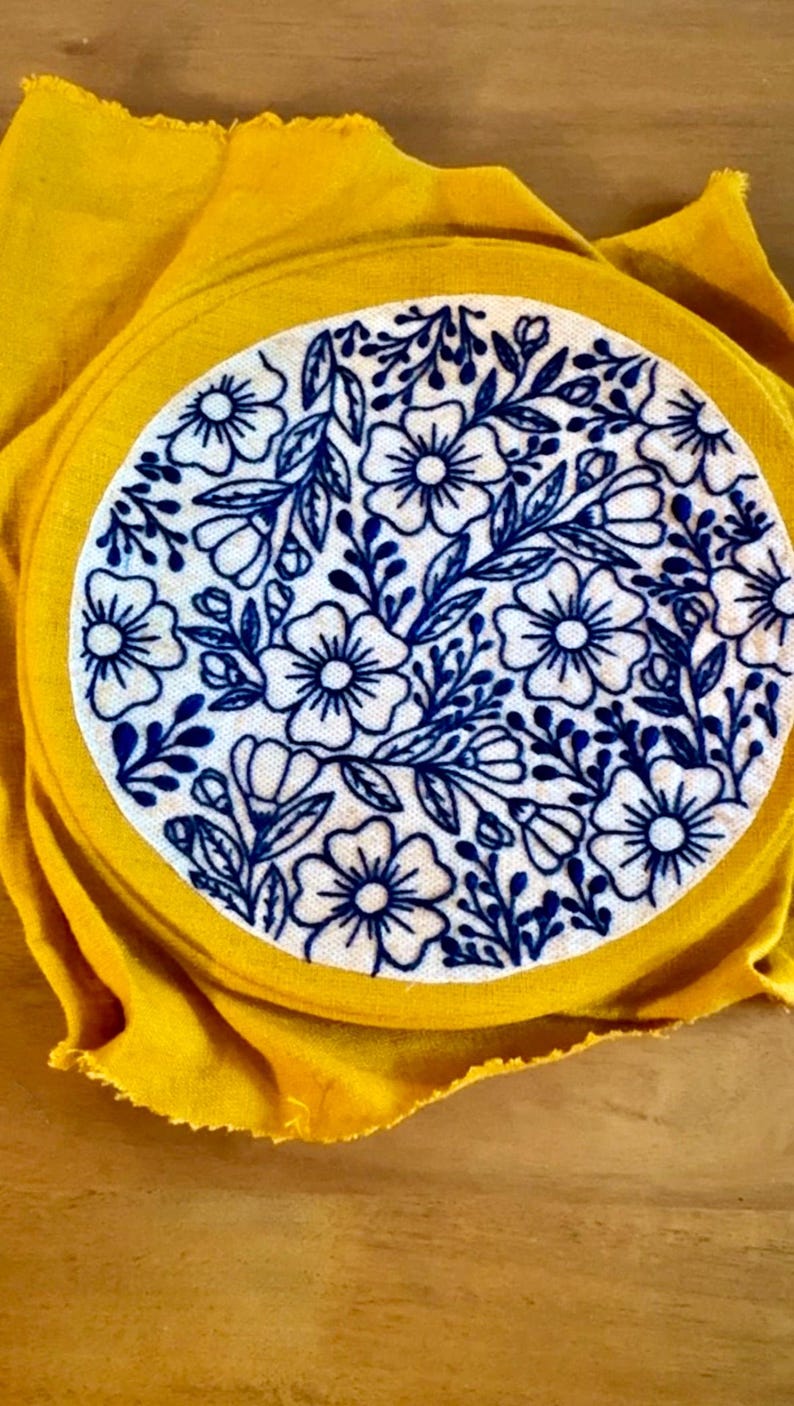Puede incluir: Un c&iacute;rculo de tela blanca con un dise&ntilde;o de bordado floral azul, sobre un fondo de tela amarilla. El bordado presenta flores y hojas detalladas. La tela amarilla rodea el c&iacute;rculo.