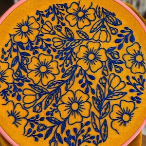 Puede incluir: Un bastidor bordado a mano con un vibrante diseño floral en hilo azul sobre tela amarillo mostaza. El bordado está sujeto en un bastidor rosa. Una pequeña etiqueta en forma de ataúd con el texto "Coffin Club" está adjunta.