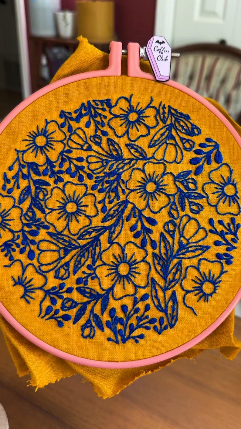 Puede incluir: Un bastidor bordado a mano con un vibrante dise&ntilde;o floral en hilo azul sobre tela amarillo mostaza. El bordado est&aacute; sujeto en un bastidor rosa. Una peque&ntilde;a etiqueta en forma de ata&uacute;d con el texto "Coffin Club" est&aacute; adjunta.