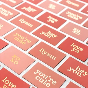 Puede incluir: Una colección de tarjetas de color rosa y melocotón con letras doradas. Las tarjetas presentan varias frases como "love ya", "you're cute", "hey you", "ilysm" y "xo xo".