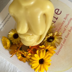 Beeswax candle in the form of a pregnant woman holding her belly is unlit on a birth altar, seen from above, surrounded by books for childbirth and calendula, an herb known for its healing support