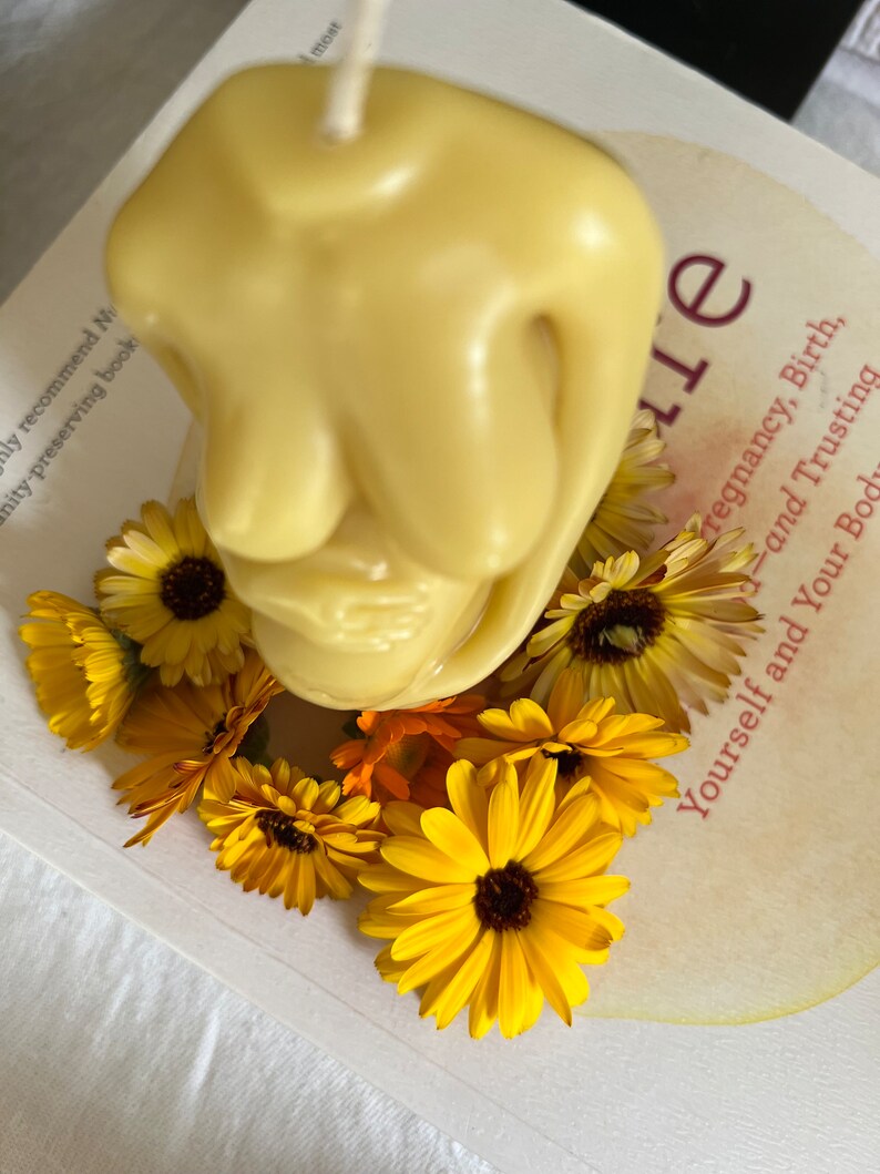 Beeswax candle in the form of a pregnant woman holding her belly is unlit on a birth altar, seen from above, surrounded by books for childbirth and calendula, an herb known for its healing support