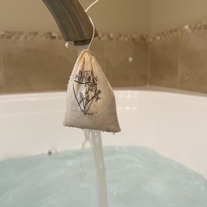 May include: A small white muslin bag with the text "Bath & Body" printed on it is being held under a silver faucet with water running into a white bathtub.