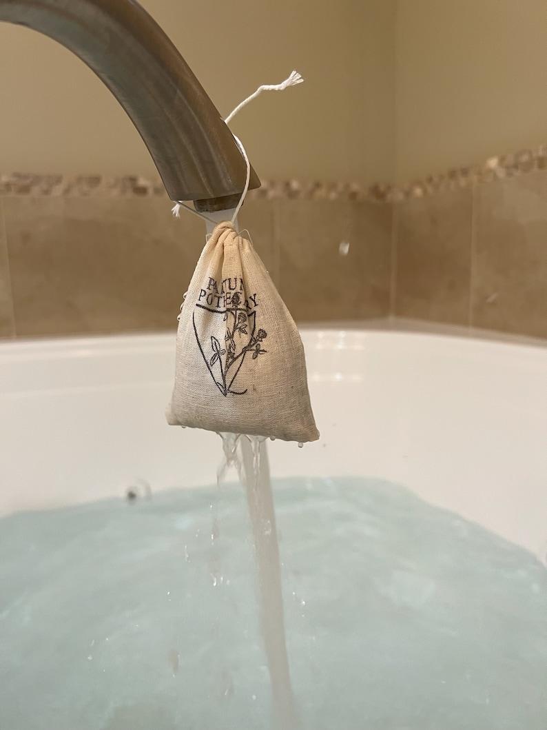 May include: A small white muslin bag with the text "Bath & Body" printed on it is being held under a silver faucet with water running into a white bathtub.