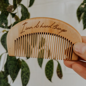 Small hand held wooden comb for natural childbirth and natural pain relief. Background environment is a houseplant in front of a white brick wall. A Mother's hand is holding the birth comb out