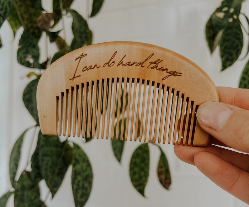 Small hand held wooden comb for natural childbirth and natural pain relief. Background environment is a houseplant in front of a white brick wall. A Mother's hand is holding the birth comb out