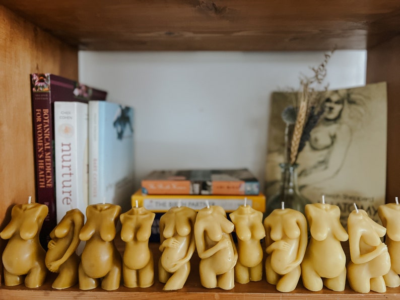 Many skin tone Beeswax candle in the form of a pregnant woman holding her belly is lit on a birth altar, surrounded by books for childbirth.Candles are displayed on a bookshelf in front of a charcoal drawing of a pregnant mom