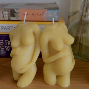 Twe beeswax candles in the mold of a pregnant woman holding her belly is unlit on a birth altar, surrounded by books for childbirth. Candle wicks are white. Unusual candle mold for moms