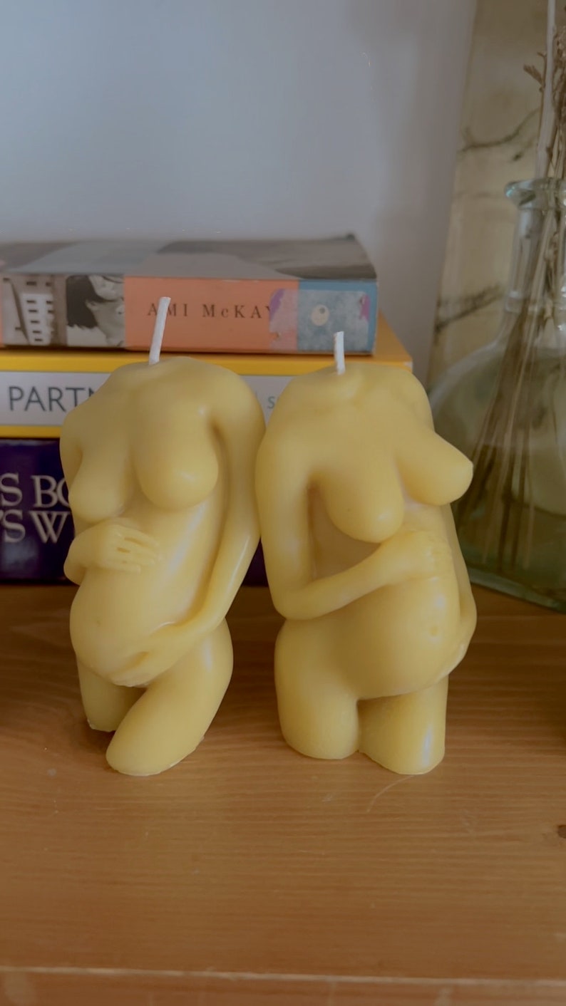 Twe beeswax candles in the mold of a pregnant woman holding her belly is unlit on a birth altar, surrounded by books for childbirth. Candle wicks are white. Unusual candle mold for moms