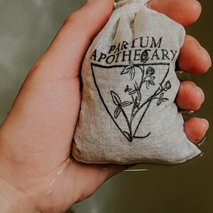 Woman's hand holds the sachet of healing herbs above a bath full of water. Sachet is stamped with partum apothecary logo of a triangle with an herbal design