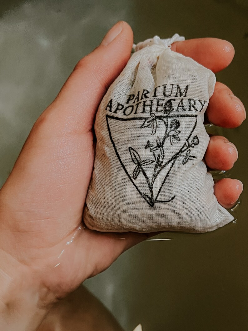 Woman's hand holds the sachet of healing herbs above a bath full of water. Sachet is stamped with partum apothecary logo of a triangle with an herbal design