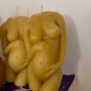 Two beeswax candles in the form of a pregnant woman holding her belly Sit on top of the book women's bodies women's wisdom. Candles cut off at the shoulders and thigh, beautiful caramel color candle