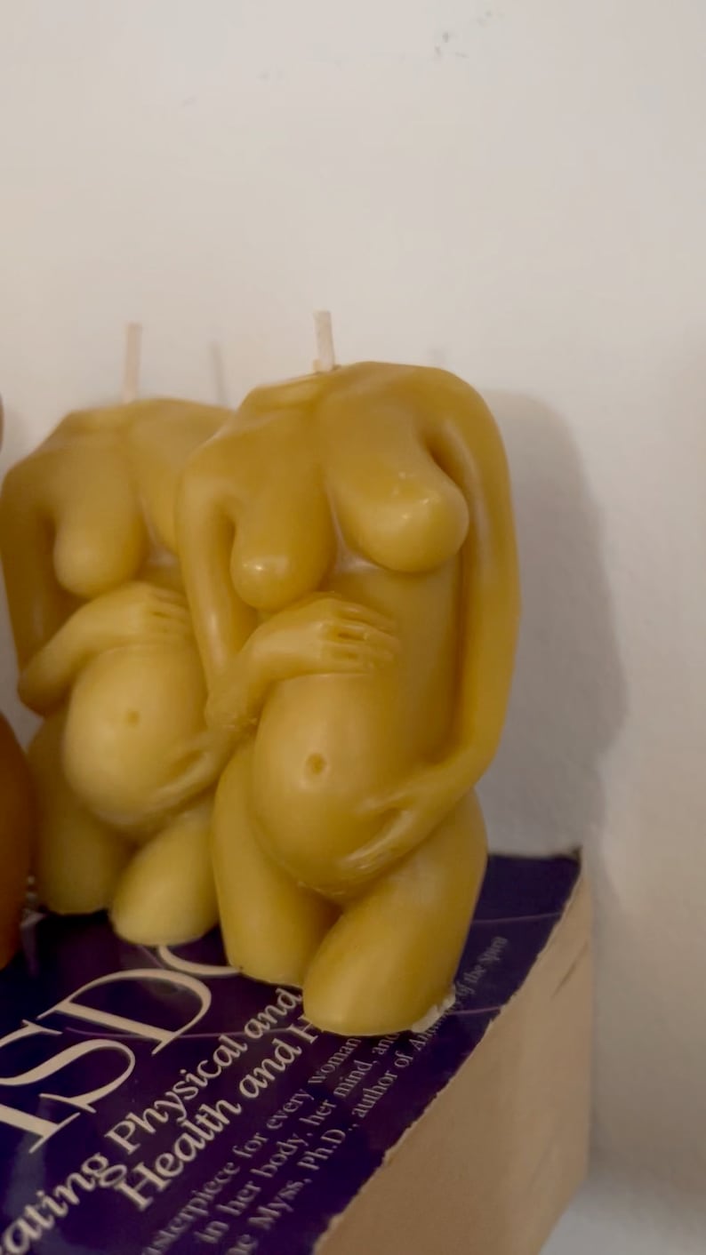 Two beeswax candles in the form of a pregnant woman holding her belly Sit on top of the book women's bodies women's wisdom. Candles cut off at the shoulders and thigh, beautiful caramel color candle