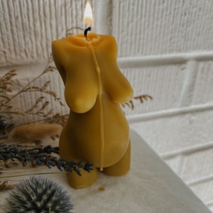 A yellow beeswax candle shaped like a pregnant belly and torso. The candle is lit and dripping wax. The candle is sitting on a white surface.