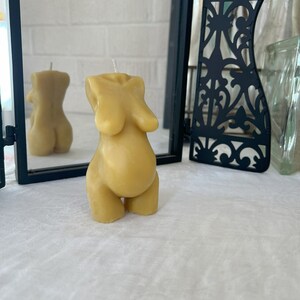 Beeswax candle in the form of a pregnant woman with big belly is lit on a birth altar, surrounded by dried healing herbs like rose and chamomile. Shows back view of the candle in the form of a woman's body. Beautiful caramel color candle