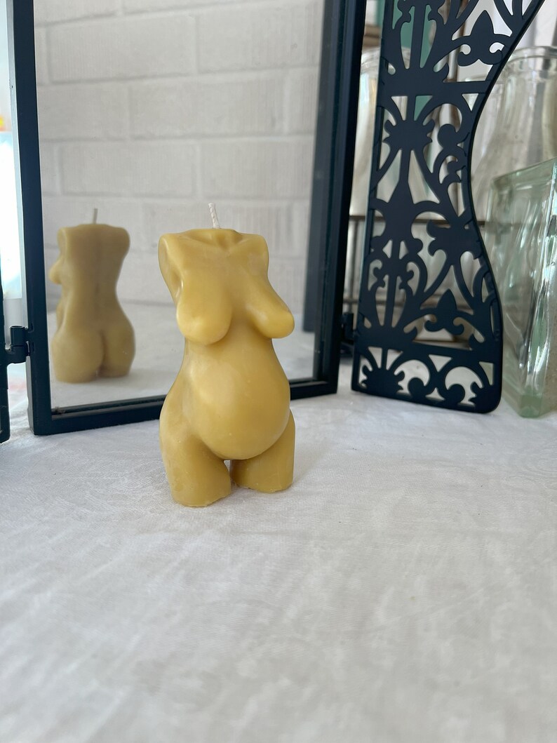Beeswax candle in the form of a pregnant woman with big belly is lit on a birth altar, surrounded by dried healing herbs like rose and chamomile. Shows back view of the candle in the form of a woman's body. Beautiful caramel color candle