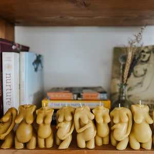 Many skin tone Beeswax candle in the form of a pregnant woman holding her belly is lit on a birth altar, surrounded by books for childbirth.Candles are displayed on a bookshelf in front of a charcoal drawing of a pregnant mom