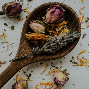 Whole herbs on a wooden spoon with scattered dried flowers. Herbs include lavender, rose and calendula, all known for healing the new mom after labor